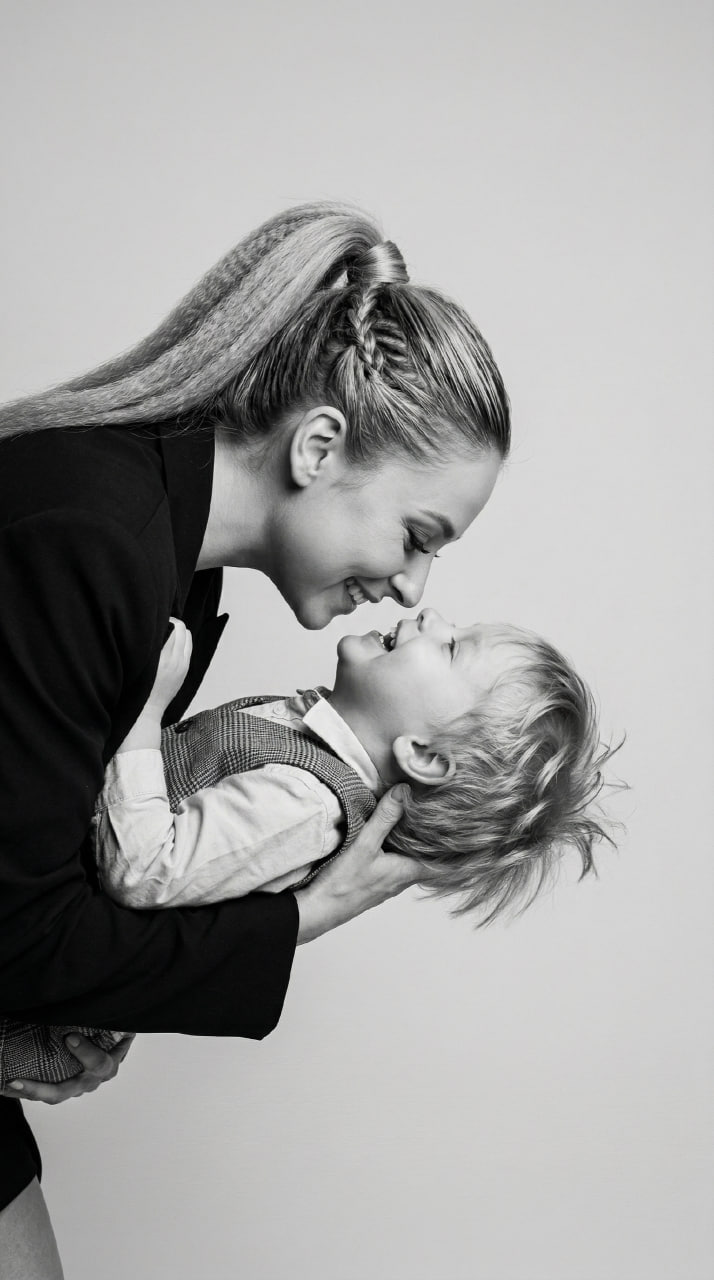 ИИ-фотосессия по фото: черно-белый студийный портрет улыбающейся мамы в темном пиджаке, бережно держащей на руках смеющегося маленького сына в жилетке.