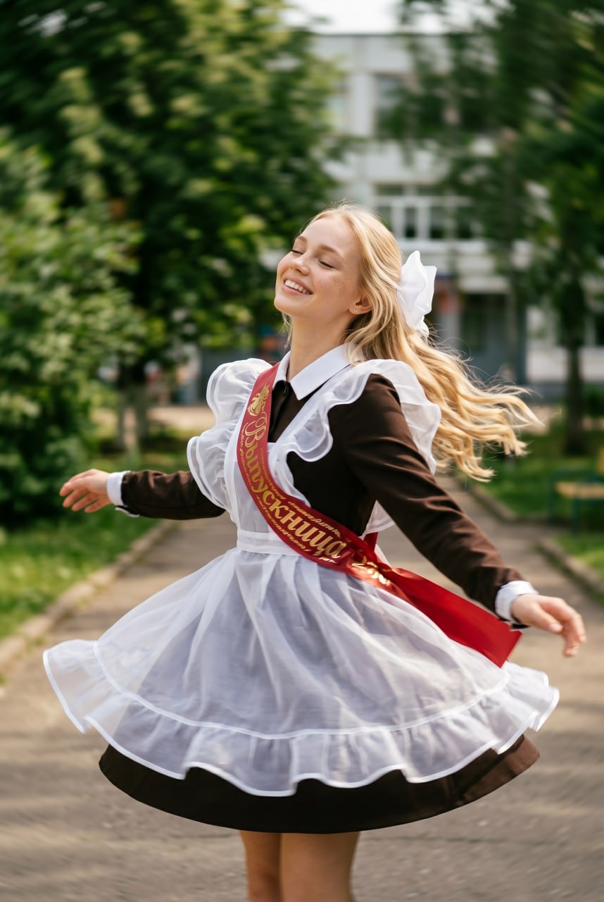 ИИ-фотосессия по фото: девушка-выпускница в классической школьной форме с белым фартуком и красной лентой кружится на школьном дворе.