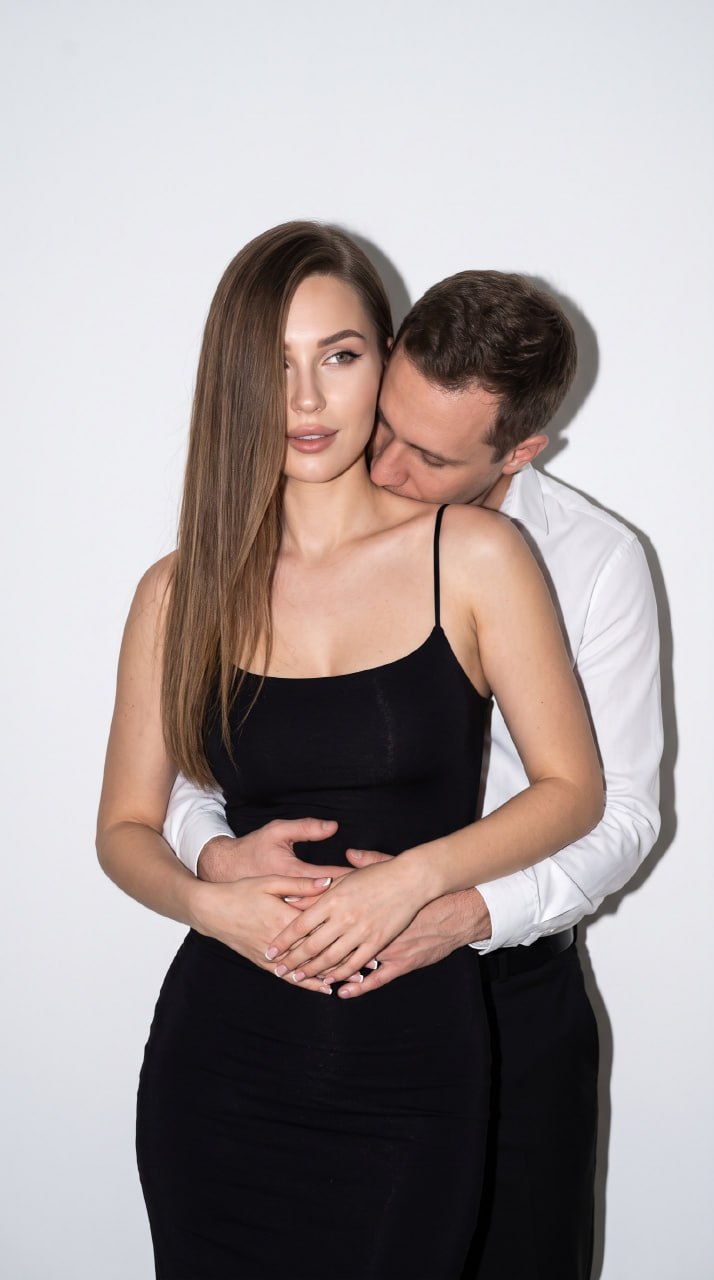 ИИ-фотосессия по фото: мужчина обнимает девушку со спины на фоне белой стены при ярком свете вспышки с четкими тенями.