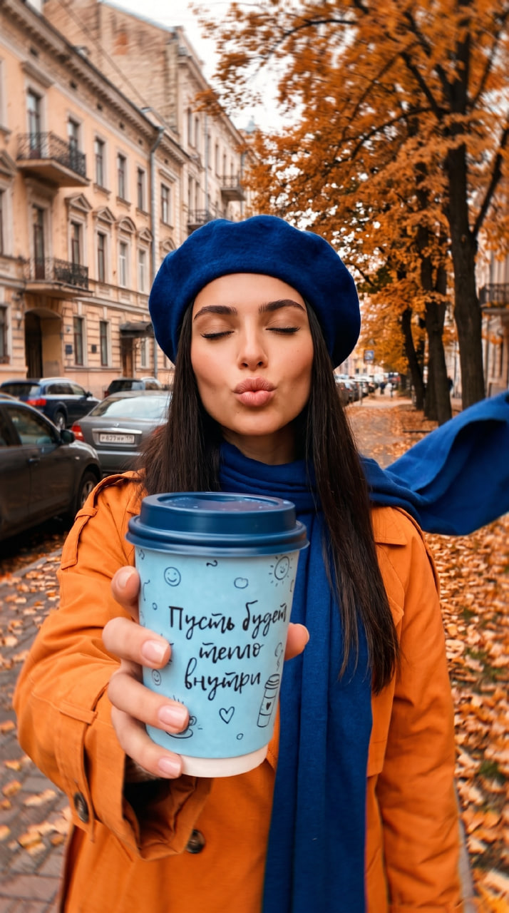 ИИ-фотосессия по фото: девушка в синем берете и рыжем тренче посылает воздушный поцелуй на осенней улице, протягивая голубой стакан кофе.