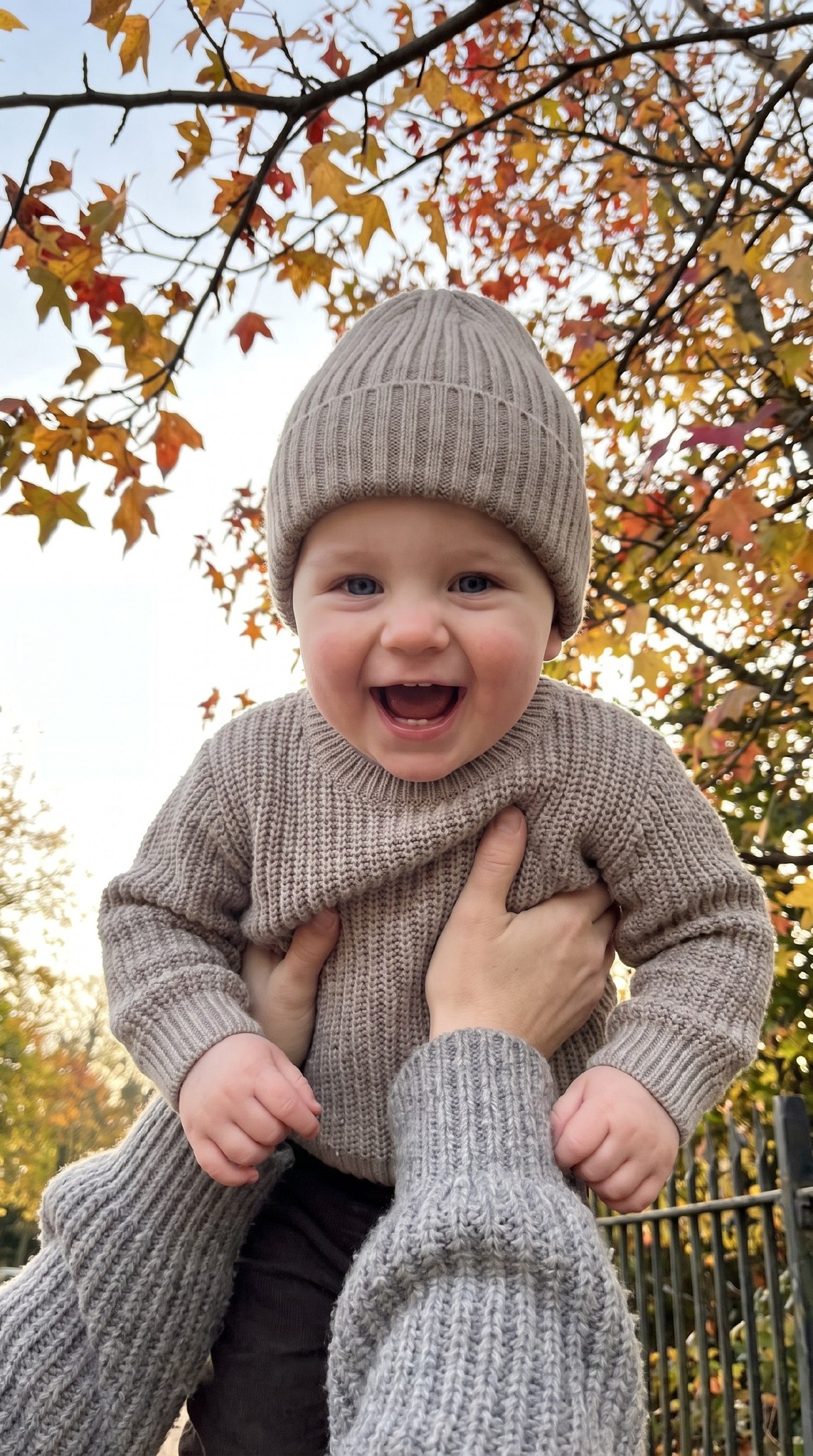 ИИ-фотосессия по фото: малыш в бежевом вязаном свитере и ребристой шапке радостно смеется на руках у взрослого на фоне осенних деревьев с золотой листвой.