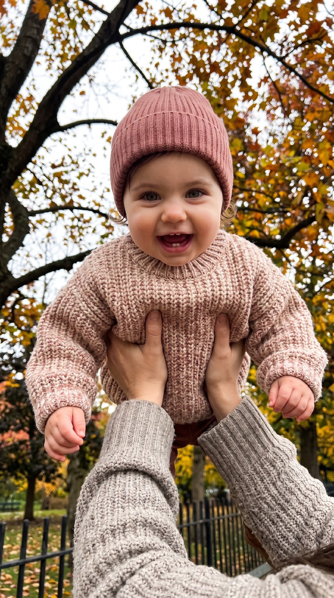 ИИ-фотосессия по фото: малыш в розовом вязаном свитере и шапке радостно смеется на руках взрослого на фоне золотых осенних крон деревьев.